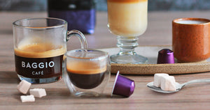 Algumas xícaras de café Baggio em uma mesa, ao lado de duas cápsulas de café roxas e uma colher com torrões de açúcar.